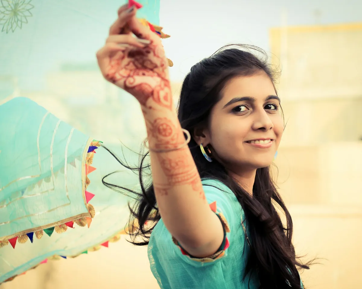 A smiling woman in a turquoise kurta, her hand painted with henna, holding up a sheer dupatta